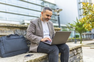 Happy mature caucasian businessman using laptop sitting on a stone bench outdoors in the financial