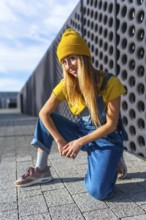 Blonde dancer wearing yellow beanie, yellow t shirt and blue dungarees, kneeling and posing in a