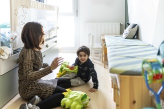Mother and son are sitting on the floor, playing with a dinosaur costume in a brightly lit bedroom