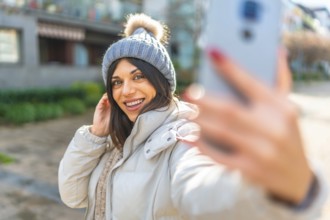 Beauty woman smiling while taking selfie with smartphone standing outdoors in a sun winter day