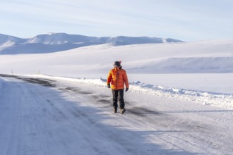 A man in an orange jacket walks across a snow-covered road. The scene is peaceful and serene, with