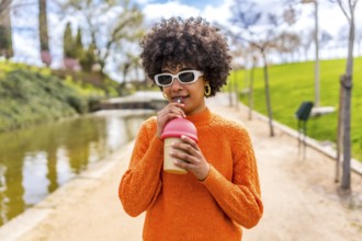 Stylish young woman with an afro hairstyle sips a beverage through a straw in a serene park setting