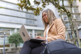 Side view low angle of a latin confident businesswoman using laptop sitting in city bench