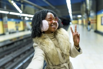 Smiling young black woman with earmuffs and sunglasses making victory sign waiting subway transport