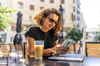 Happy arab woman using mobile app and working with laptop in an outdoor cafeteria