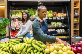 Grocery store employee arranging fresh fruit while holding a tablet, with a customer and shelves