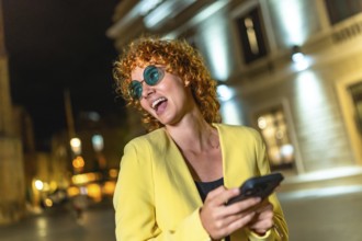 Modern woman in colorful casual clothes and blue sunglasses using smartphone laughing in the city