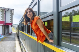 A woman in an orange shirt is leaning out of a train window. The train is parked at a station