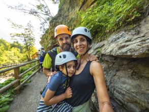 Family wearing helmets taking selfie while hiking on wooden walkway next to rock wall in beautiful