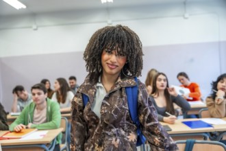 Confident young university student standing in classroom with backpack, classmates studying in