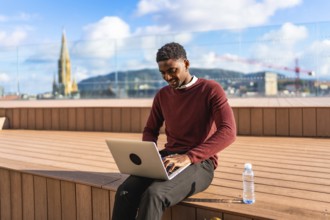 African male young entrepreneur using laptop sitting on a city terrace under the sun