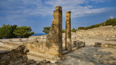 Two ancient stone columns stand in the middle of a ruined site under a slightly cloudy sky,
