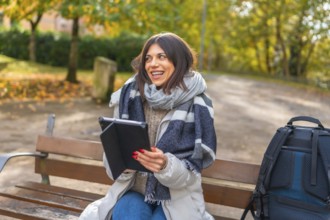 Happy caucasian female student using digital tablet sitting on a park bench during a cold day of