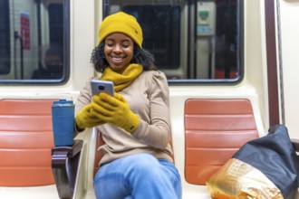 Smiling young woman wearing yellow gloves and beanie using smartphone while commuting on subway