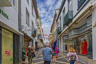 People walk through a busy shopping street with modern shops and historic buildings under a bright