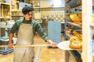 Latin young man using a wooden peel to remove baking bread from traditional oven