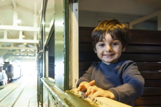 A young boy is smiling and looking out a window of a train. The train is in a station and the boy