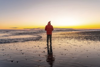 A man stands on a beach at sunset, wearing a red jacket. The scene is serene and peaceful, with the