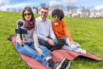 Three content creators sitting on a blanket in a park, recording a video blog using a smartphone