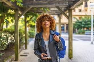 Latin beauty woman with curly hair using phone while walking along a city carrying rucksack