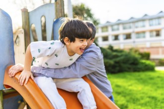 Grandfather holding grandson sliding down an orange slide at playground, having fun together on a