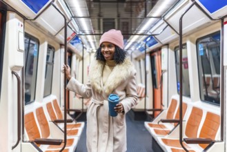Smiling young black woman wearing winter coat and pink woolen hat holding reusable coffee cup