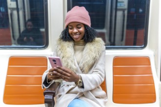 Smiling young black woman wearing winter clothes using smartphone while sitting on subway train