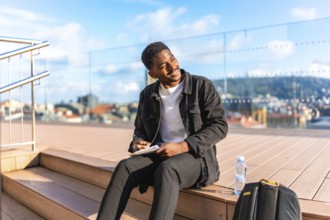 African man writing poems by hand in a notebook sitting on a city rooftop