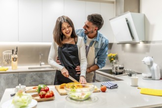 Young smiling couple wearing aprons preparing a healthy vegetable salad together in their modern