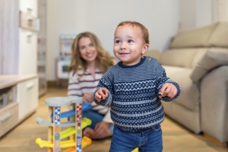 Happy baby boy learning to walk in living room with his mother watching and smiling, playing with a