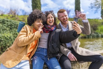 Three cheerful multi ethnic friends are making a selfie while sitting on a wall in a park next to a