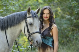 A woman in a brown and blue dress stands close to a Quarab horse in a peaceful, wooded area,