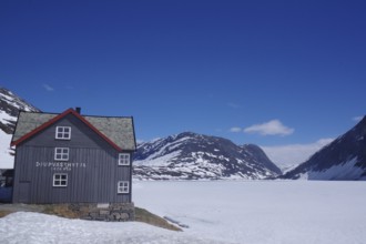 Mountain hut in a snow-covered late winter landscape, surrounded by mountains and a frozen lake,