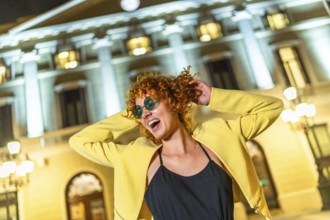 Cool happy caucasian woman in yellow jacket and sunglasses dancing in the city at night