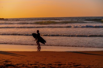 A man is walking on the beach with a surfboard. The sky is orange and the water is calm