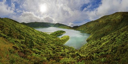 A clear lake surrounded by green hills and mountains under a blue sky with clouds on a sunny day,