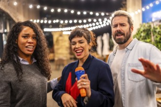 Multi-ethnic happy friends complain while woman singing karaoke at night party in a bar terrace