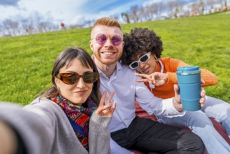 Three happy multi ethnic friends are taking a selfie in a park, gesturing victory signs and showing