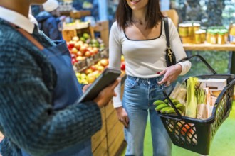 Saleswoman using digital tablet assisting customer carrying shopping basket with fresh produce in