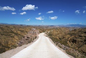 A long straight road leads through a vast desert landscape with mountains and a clear blue sky,
