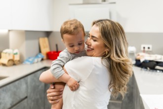 Smiling mother holding her baby son in a home kitchen, enjoying a tender moment of affection and