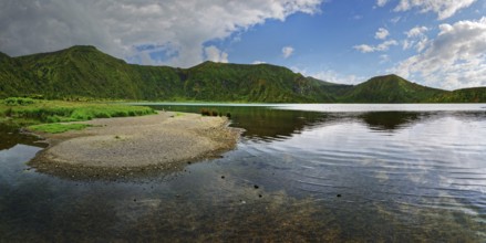 Calm lake surrounded by green mountains under a partly cloudy sky, crater lake Lagoa do Fogo,