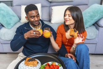 Multiethnic couple sits together, enjoying breakfast and wine, creating a cozy and intimate dining