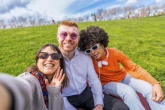 Three multi ethnic friends are sitting on the lawn in a park, taking a selfie and waving at the