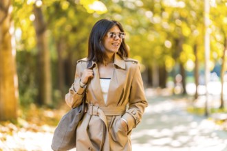 A woman wearing a tan trench coat and sunglasses walks down a sidewalk. She is smiling and she is