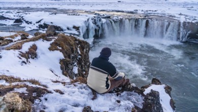 A man sitting looking at the frozen Godafoss waterfall at sunset in winter, Iceland, Europe