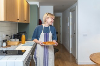 Smiling woman wearing apron carrying plate of fresh croissants in contemporary kitchen, enjoying