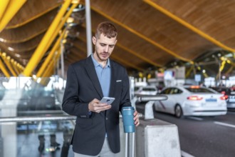 Young businessman using smartphone and holding reusable coffee cup while waiting for taxi or