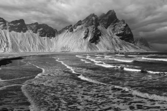 White foamy waves on the beach of Stokkness, behind snowy rocky slopes of Vestrahorn, black and