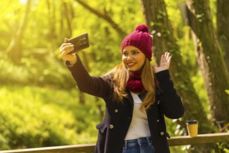 Young man in a black jacket, scarf and red woolen hat enjoying in an autumn park, waving on a video